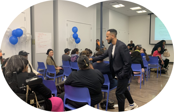 A community event in a modern meeting room with rows of blue chairs. People are seated and engaged in conversation, while a person in a suit interacts with attendees. Blue and white balloons decorate the space, and a presentation screen is visible in the background.